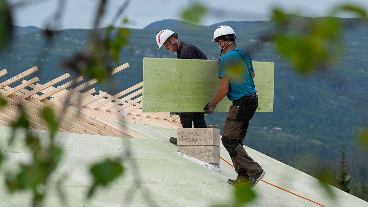 To menn som monterer takplater på hustak. Den ene bærer en kontruksjonsfinerplate.